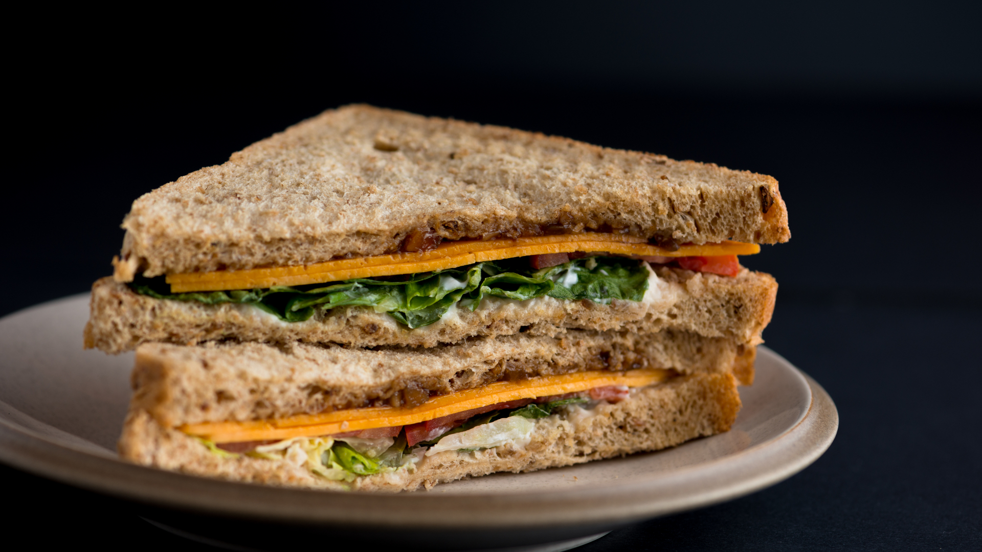 A plated cheese and salad sandwich with mayonnaise on brown bread against a black background.