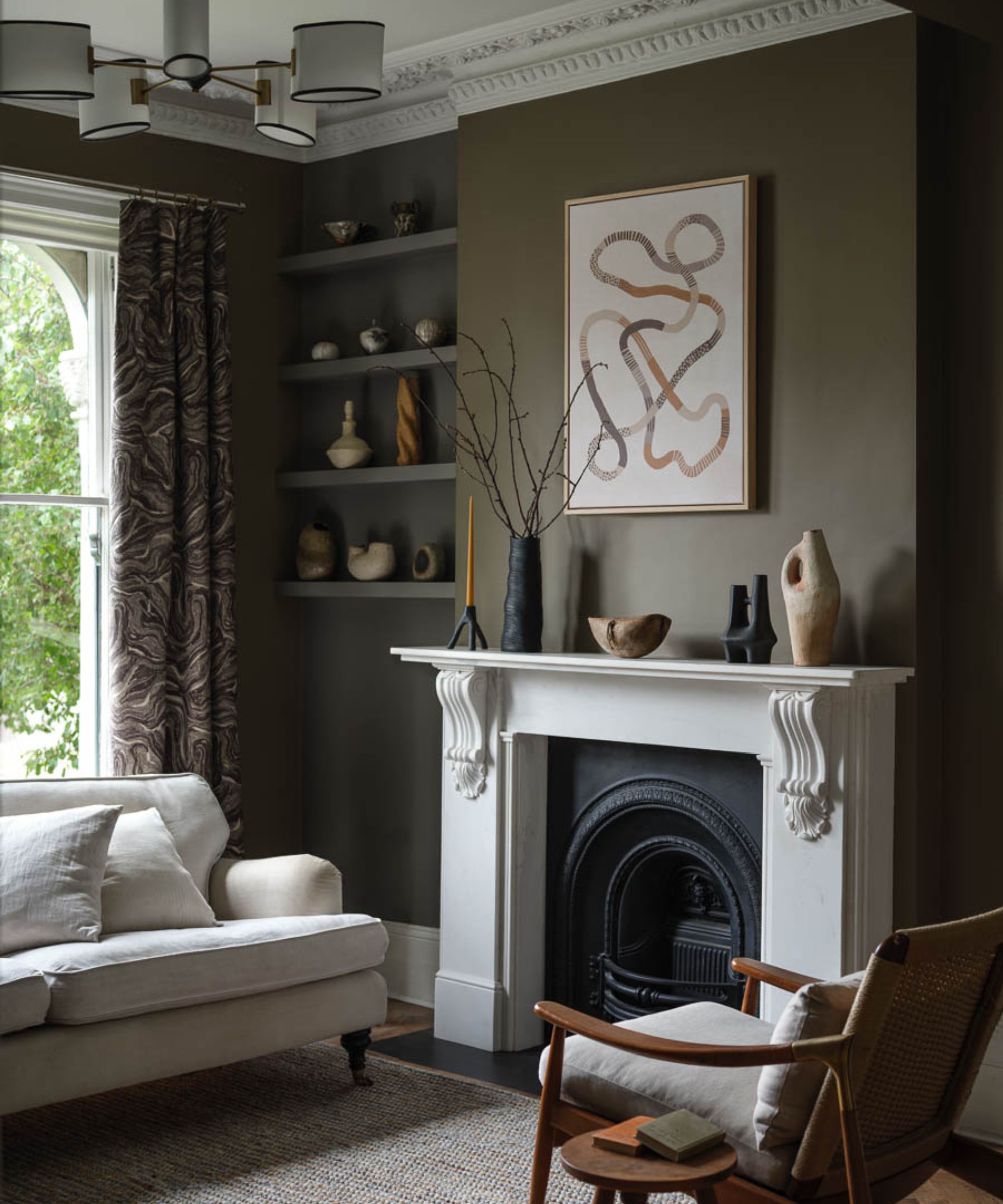 Living room in a Victorian home with a large white and black fireplace, dark olive green walls, a wooden lounge chair, a cream linen sofa, abstract ceramics on the mantelpiece and shelves.