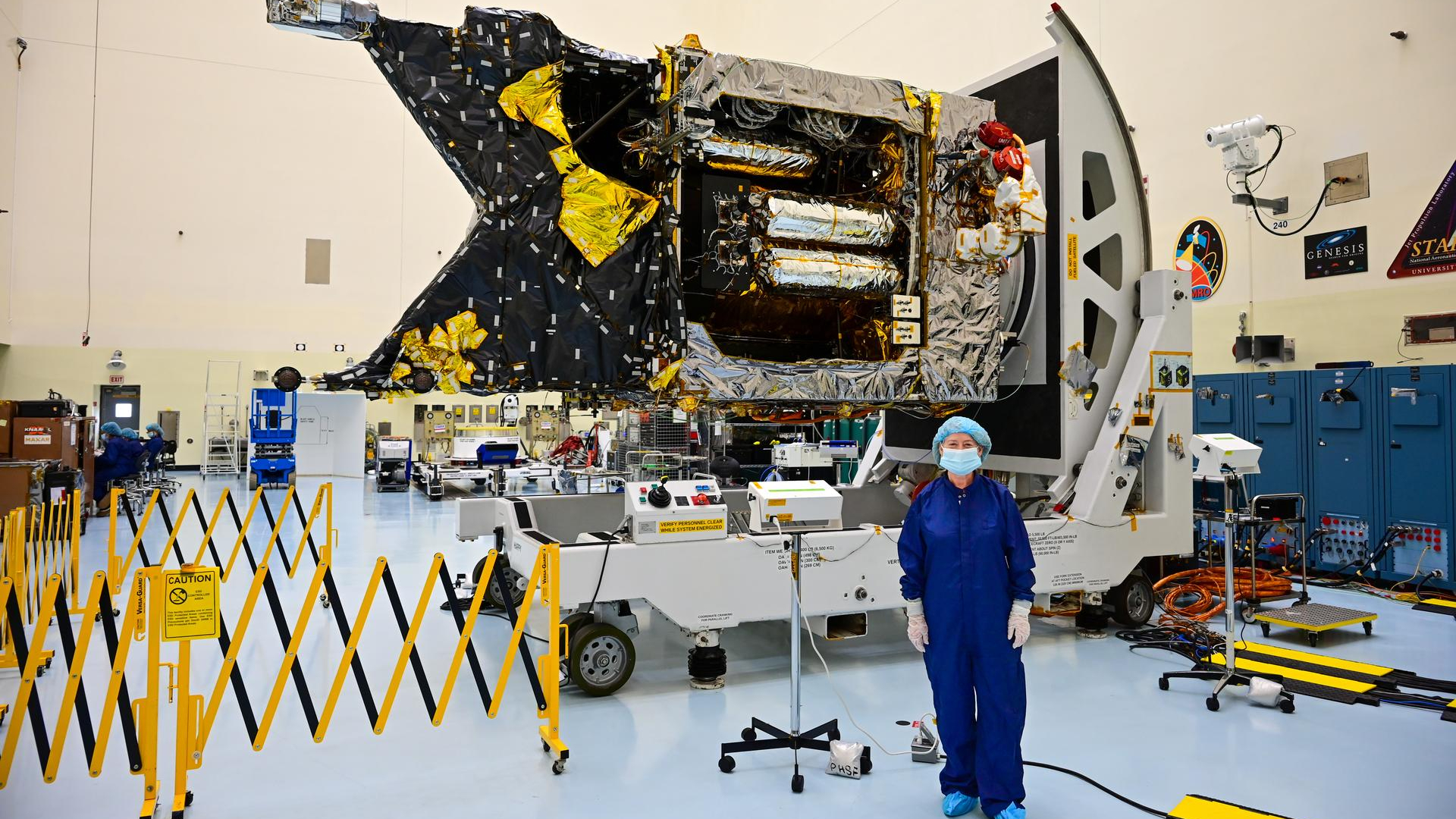 Pam Melroy stands infront of the Psyche spacecraft wearing blue overalls, blue covers over her shoes, a facemask and protective gloves.