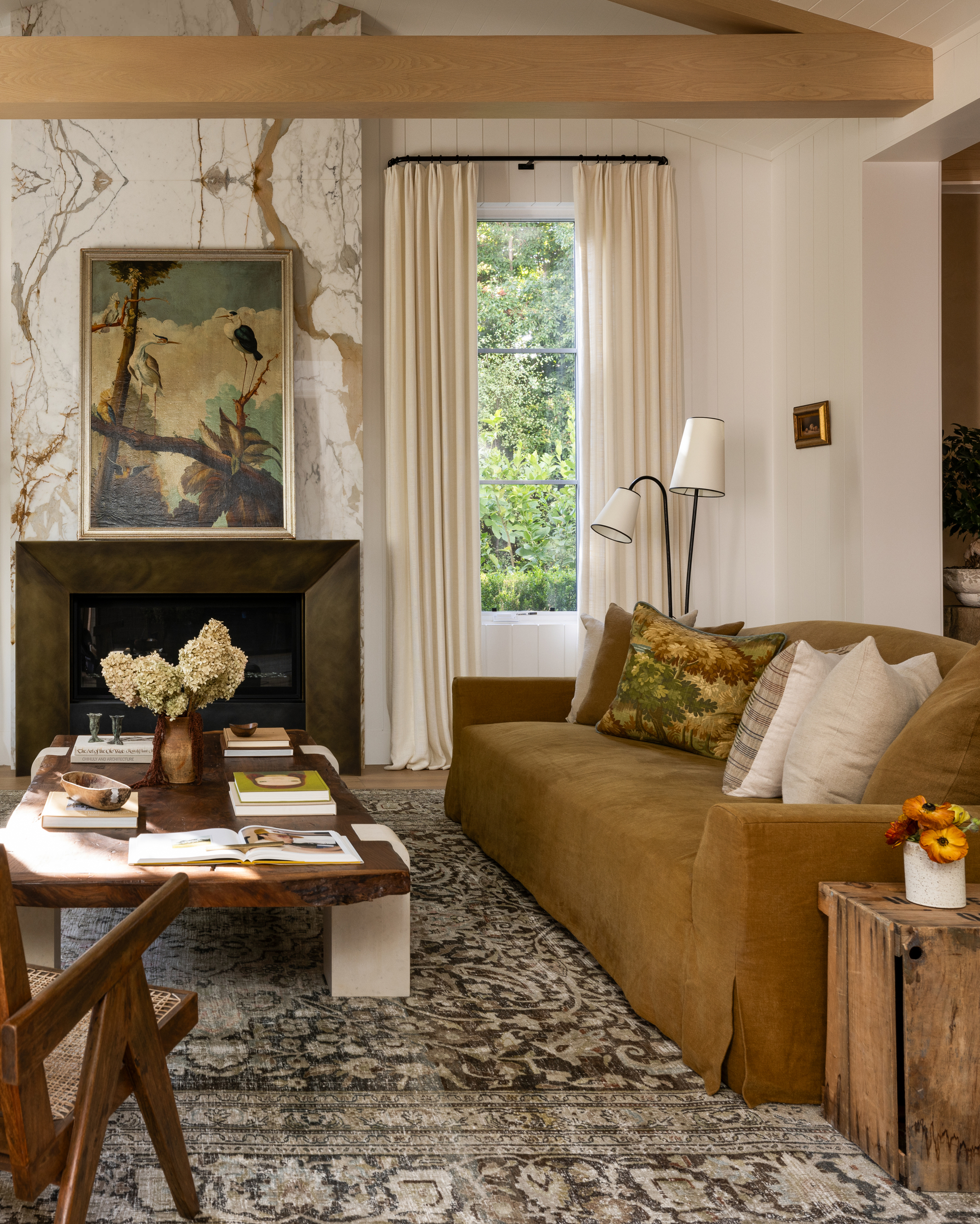 living room with patterned rug, ochre slipcover sofa with cushion, timber coffee table, cream curtains, fireplace with wallpapered wall, and exposed ceiling beam