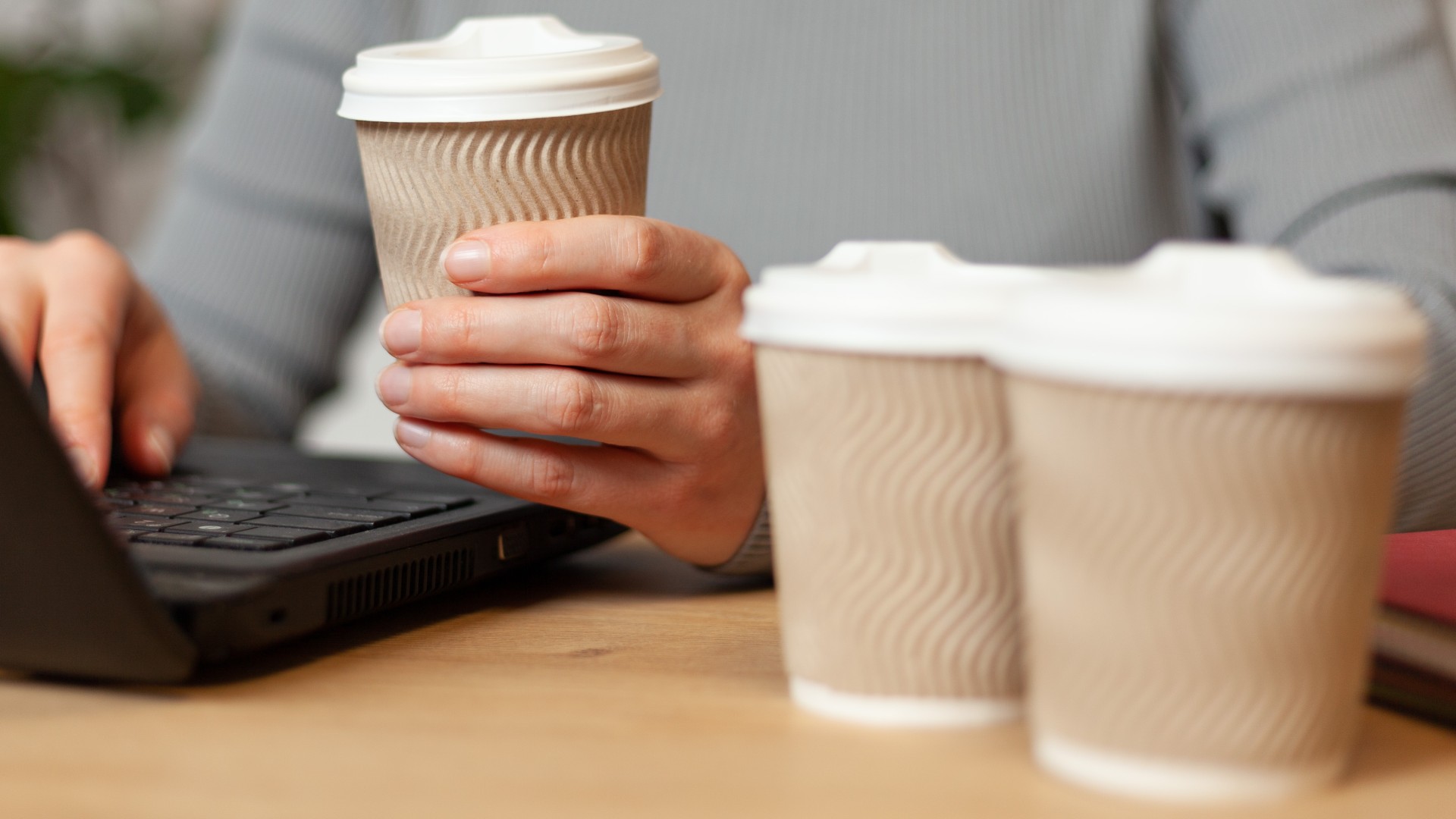 Close-up of a person working on a laptop and drinking coffee from a paper cup, with two more paper cups next to it.