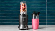 A nutribullet personal blender with fruit inside, beside a container with pink smoothie inside, against a blue subway tile wall.