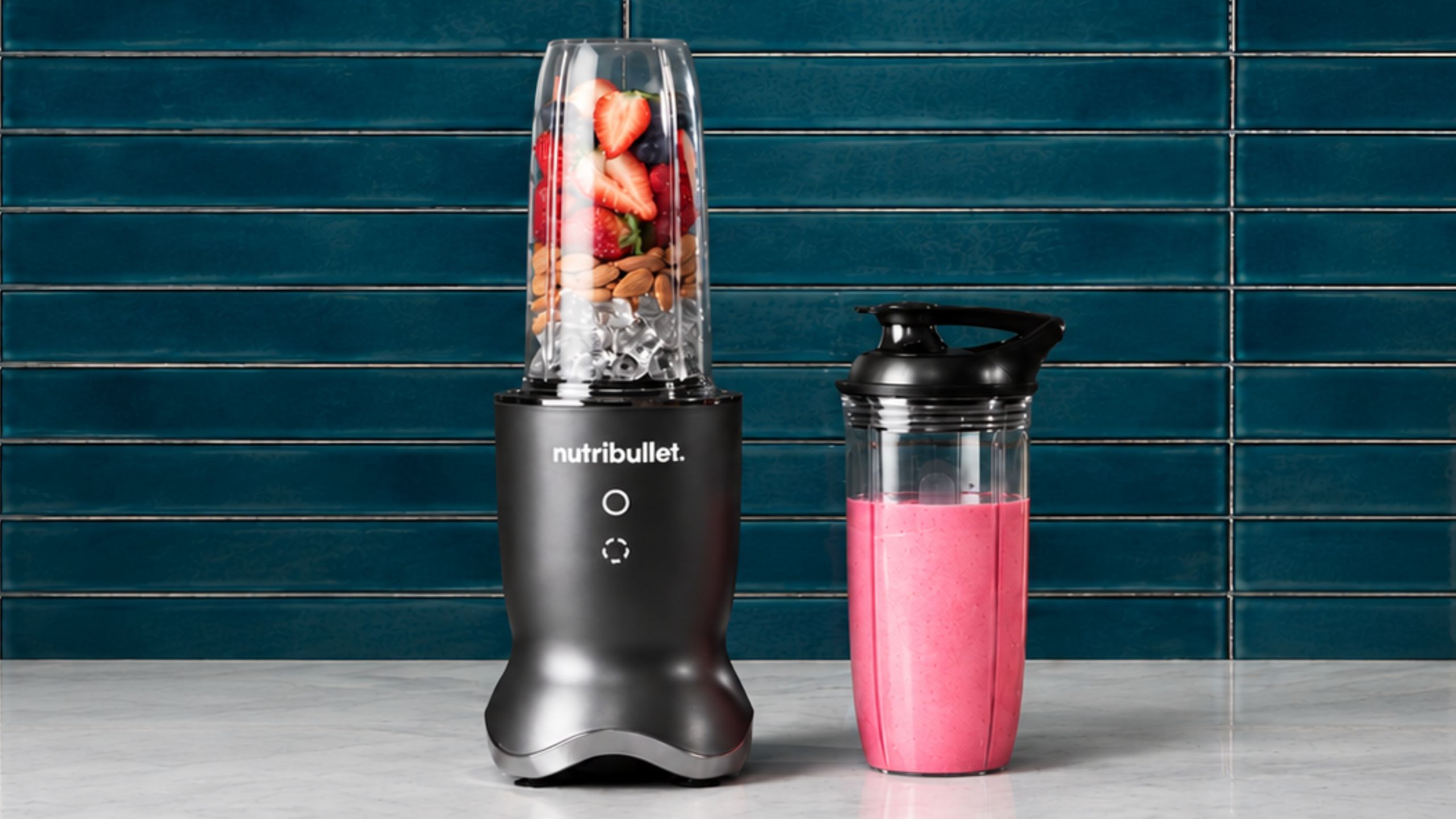A nutribullet personal blender with fruit inside, beside a container with pink smoothie inside, against a blue subway tile wall.