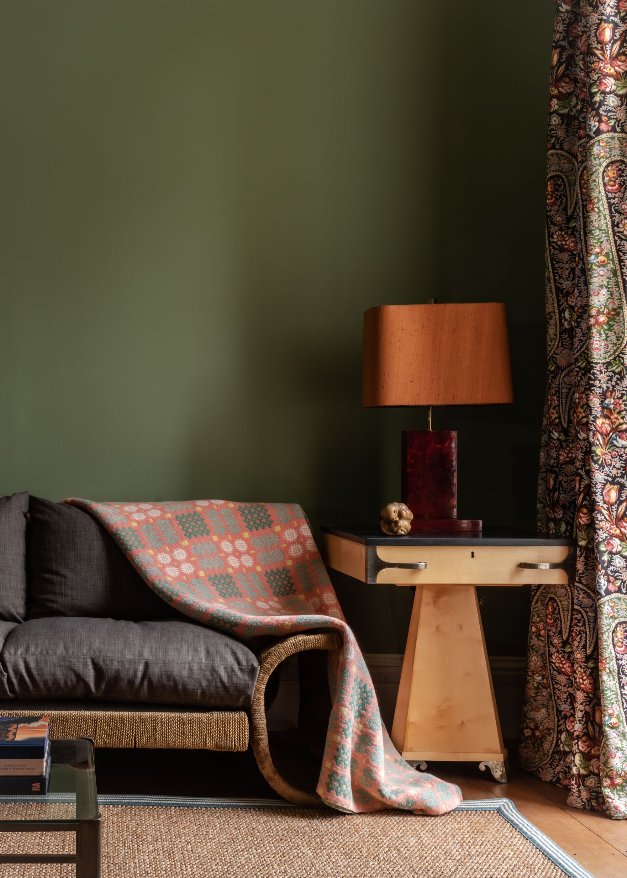 A green living room with a woven-frame sofa with gray cushions and a patterned throw blanket draped over it next to a side table with a table lamp and patterned curtains