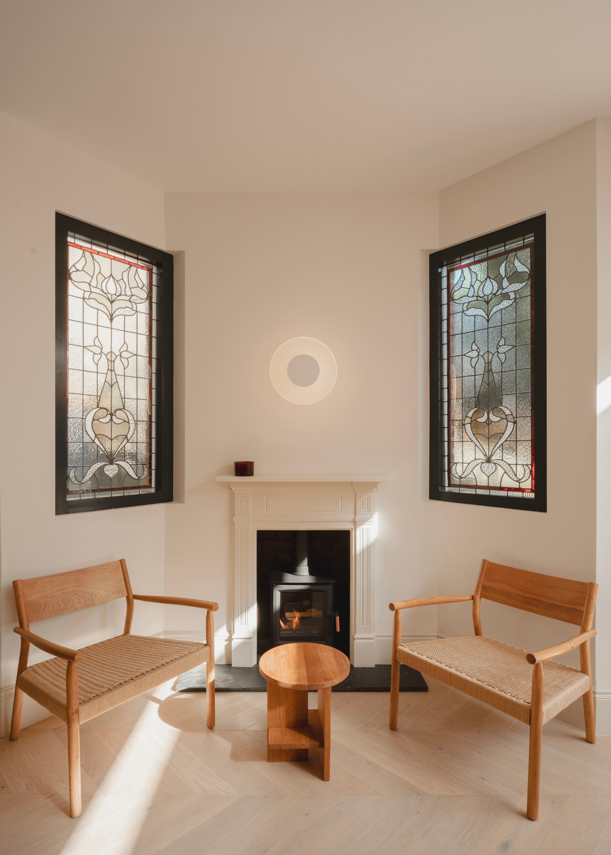 A small living space with a fireplace, a pair of wooden chairs, a small wood coffee table, a wall-mounted lamp and a pair of stained glass windows