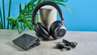 the soundcore space one pro headphones; a photograph of a pair of black over-ear headphones with cushioned cups and a soft headband, photographed in front of a blue background, showing all the accessories like USB-C charger, 3.5mm aux cable, and carry case