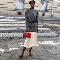 Sylvie Muse wearing a grey sweater, black belt, midi skirt, black heels, and a red Savette bag.