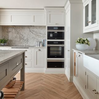 white kitchen with grey painted island, grey marble worktops and backsplash, herringbone floor boards, vase of flowers