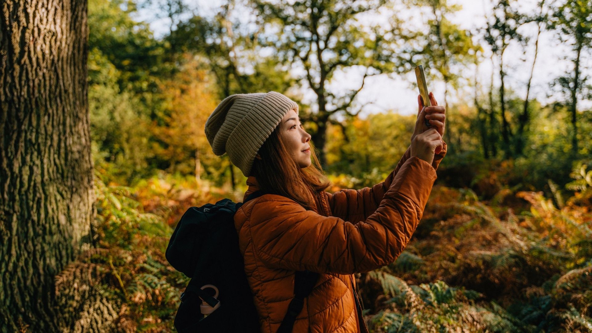 The image shows a dark haired woman wearing an orange jacket and a beige beanie taking a photo of her surrounding scenery