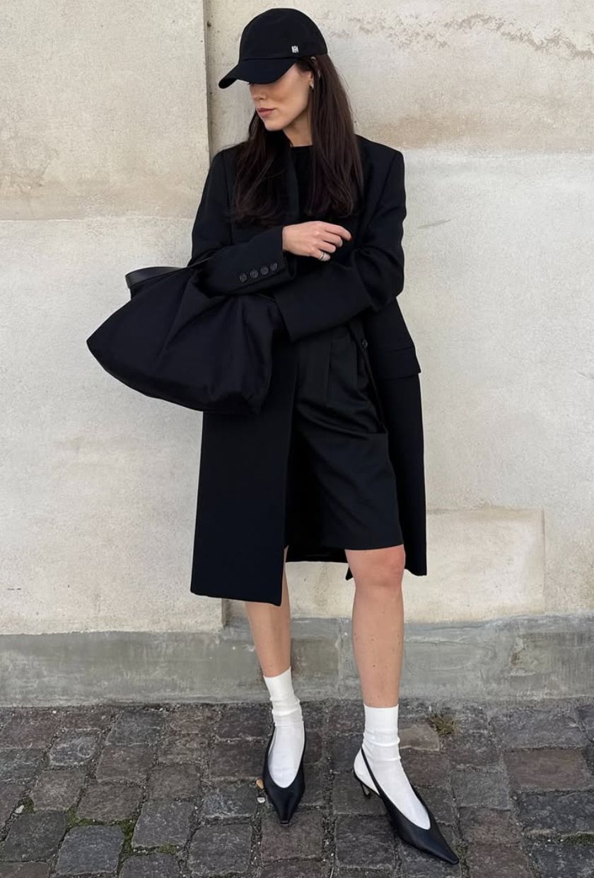@annabelrosendahl wears a long black wool coat, jumper, Bermuda shorts, white chunky socks, black slingback heels, a bag and cap