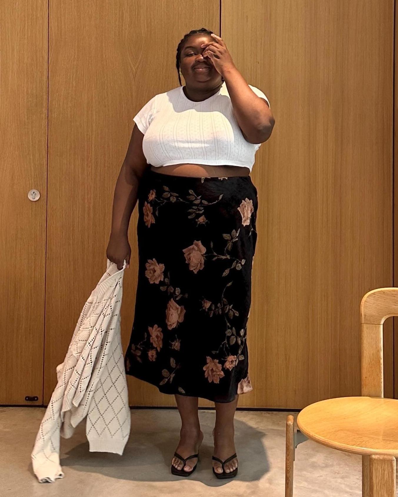 British plus-size influencer Abisola Omole wears a white cropped t-shirt, floral-print midi skirt, and black flip-flop sandals.