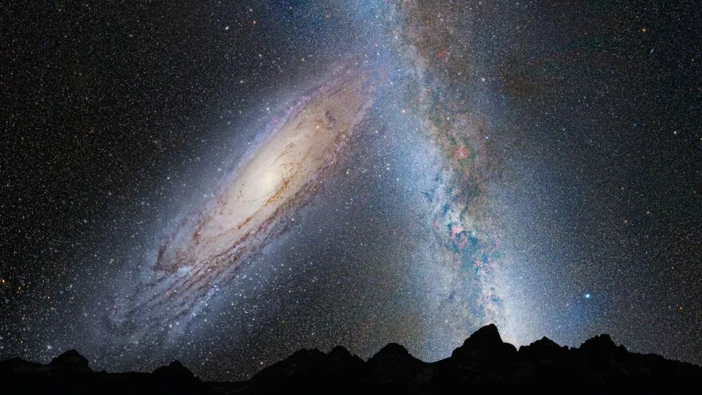 A silhouette of a mountainous horizon lays low on the image below a vast sky of stars with the arm of the milky way shooting up from the right, and a giant galaxy filling the left of the sky.