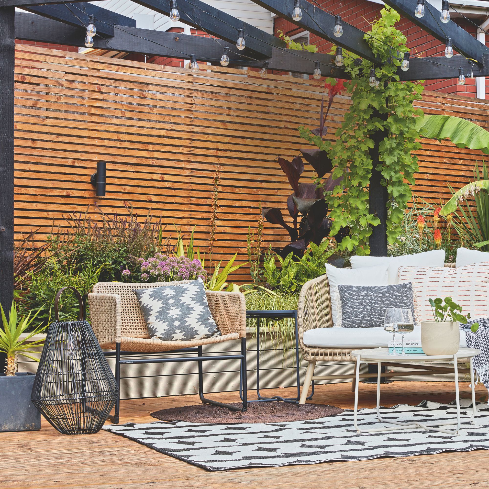 Outdoor seating area with cushions beneath wooden pergola with festoon lights and downlight on rear wood panel wall