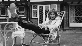 Rod Stewart in a garden, seated in a deckchair with his feet up on a table