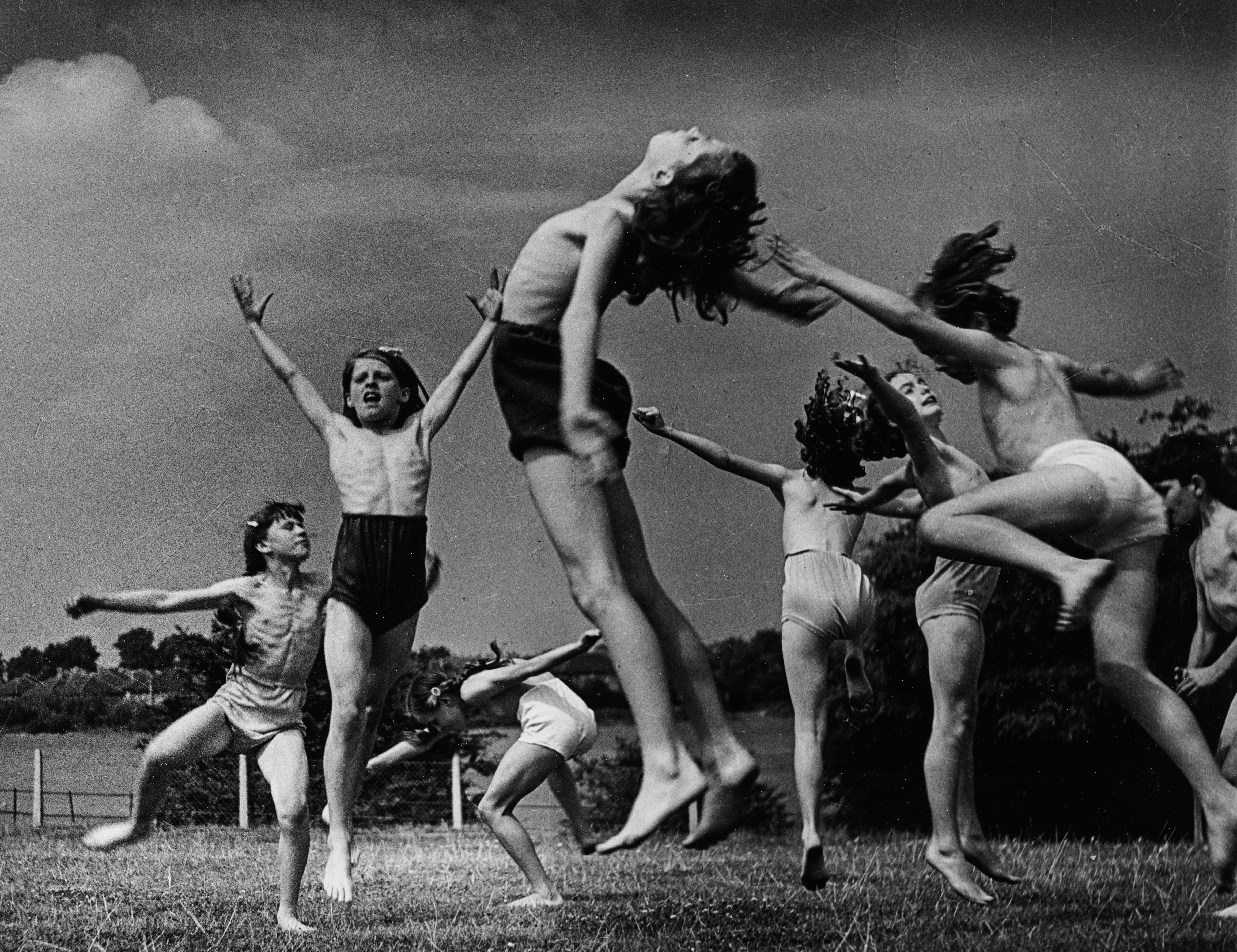 A group of children in shorts and minimal clothing leap, jump and stretch expressively on open grass, arms flung wide, bodies caught in mid-motion against a bright cloudy sky. The image conveys spontaneous physical energy and has the feel of a choreographed outdoor movement session.