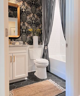 Bathroom with vanity, toilet and bath with black and white drapes, black hexagonal tile floor, jute rug, and patterned wallpaper