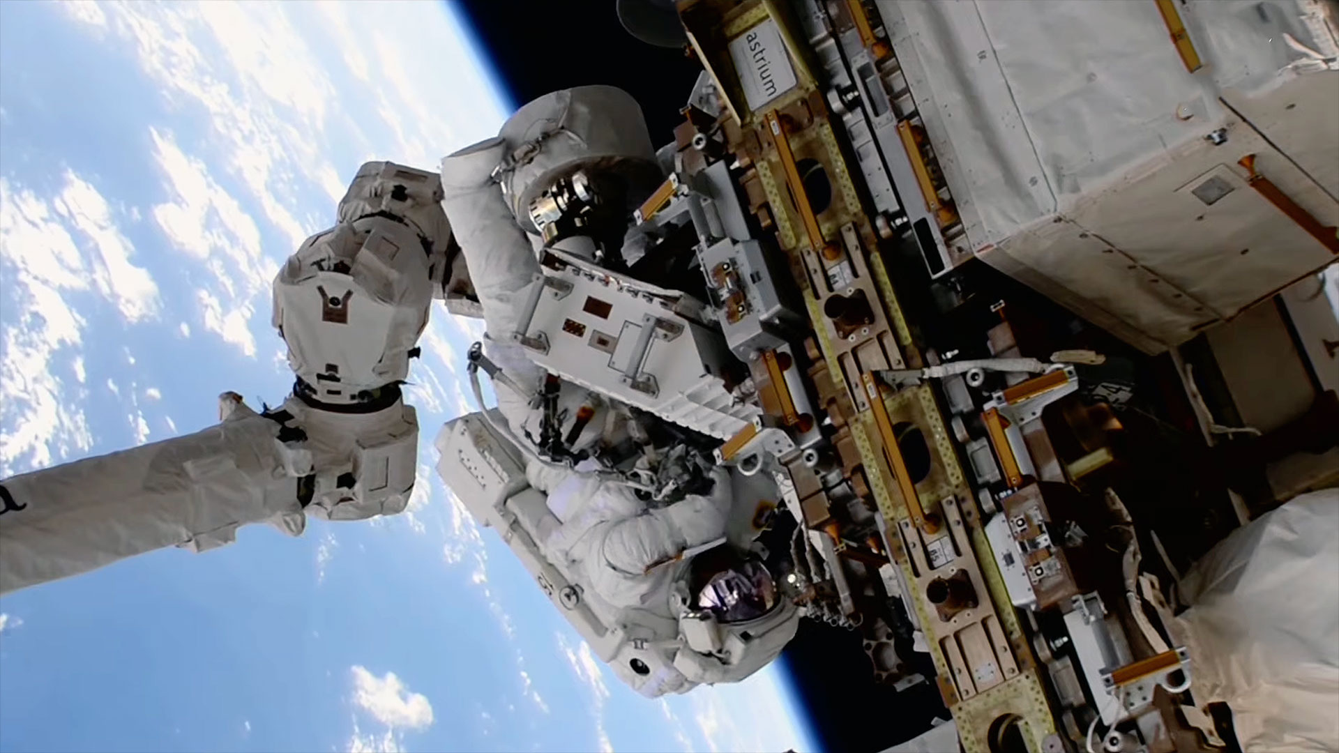 an astronaut in a white spacesuit holds a piece of equipment near the truss of a space station during a spacewalk