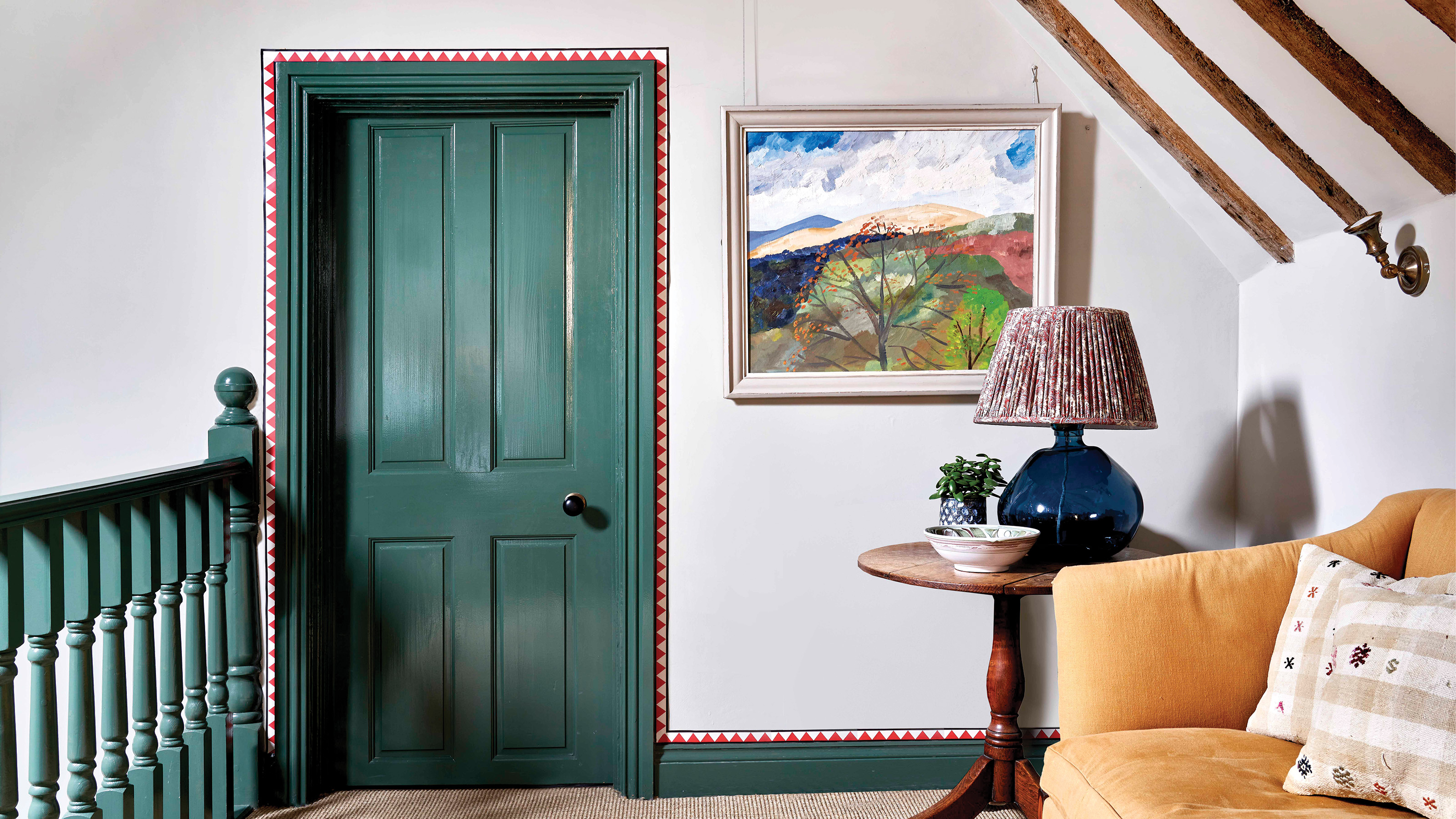 hallway with white walls and green doors and banister with red and white geometric border around the door and skirting border with natural carpet and artwork on the wall and a yellow velvet sofa to the right