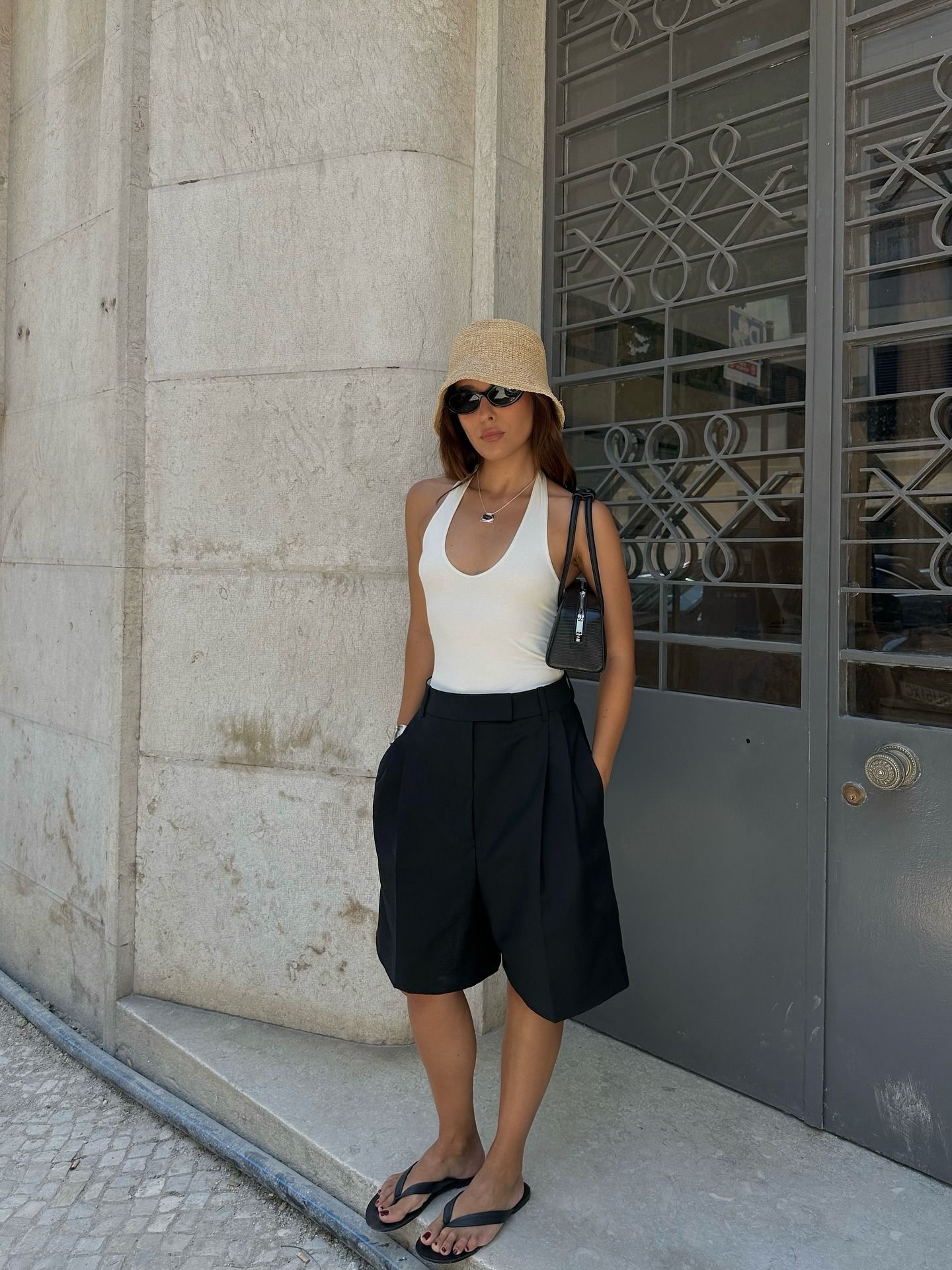 Fashion person Debora Rosa wears black Bermuda shorts, a white halterneck top crochet bucket hat, a black Alaia bag and black thong sandals in a recent image from her Instagram.