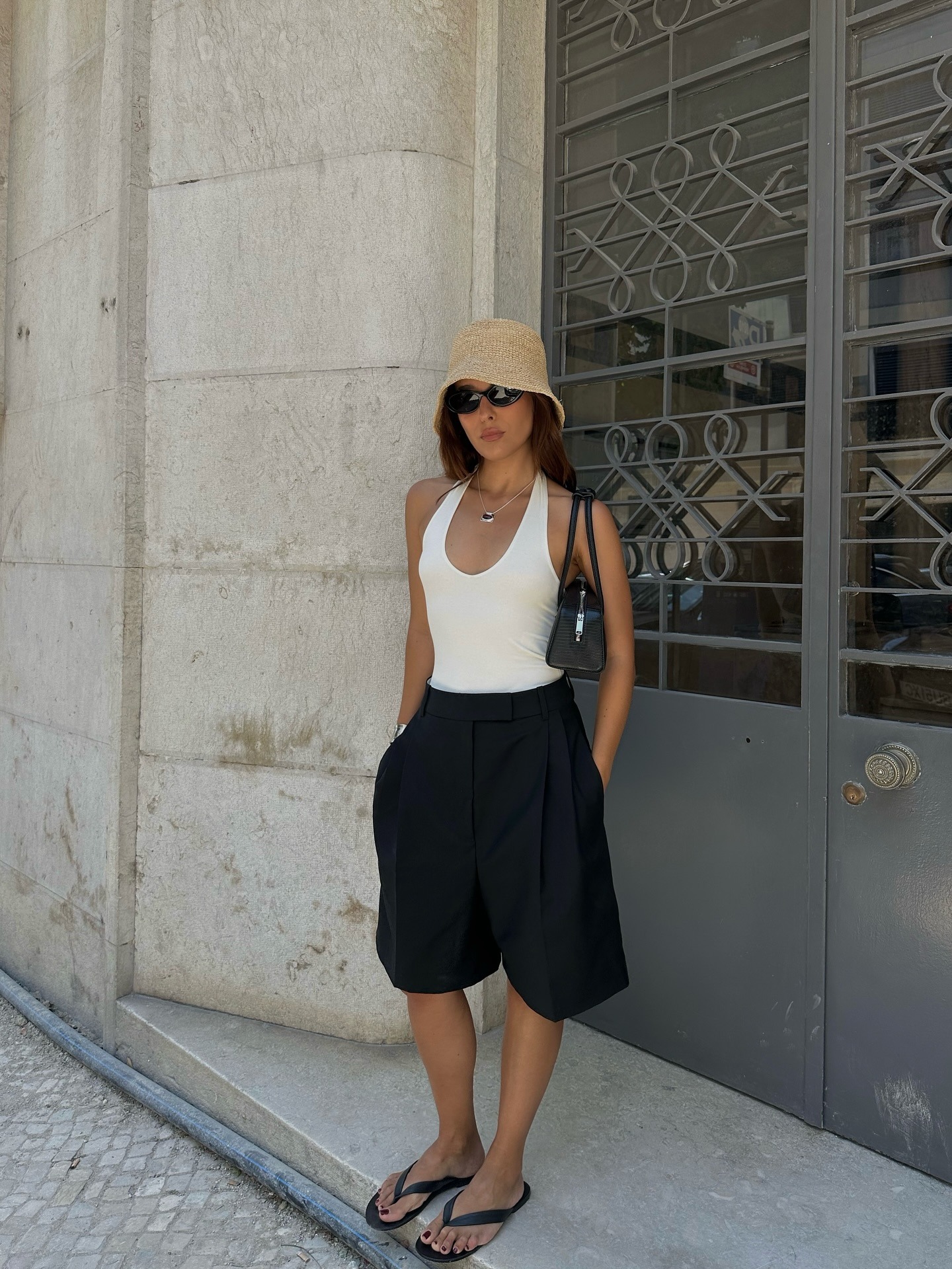 Fashion person Debora Rosa wears black Bermuda shorts, a white halterneck top crochet bucket hat, a black Alaia bag and black thong sandals in a recent image from her Instagram.