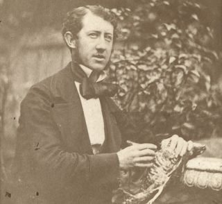 We see a black-and-white photo of a man sitting down outside and holding a dead bird.