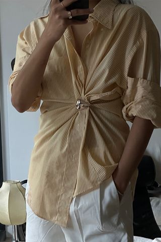 how to make a button-down shirt look more expensive is shown in a close mirror selfie photo of a woman wearing a yellow pinstripe button-down shirt bunched at the waist with a silver brooch styled with silver rings and white trousers