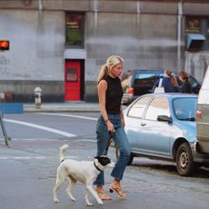 Carolyn Bessette-Kennedy wearing a black top with jeans and sandals while walking a dog. 