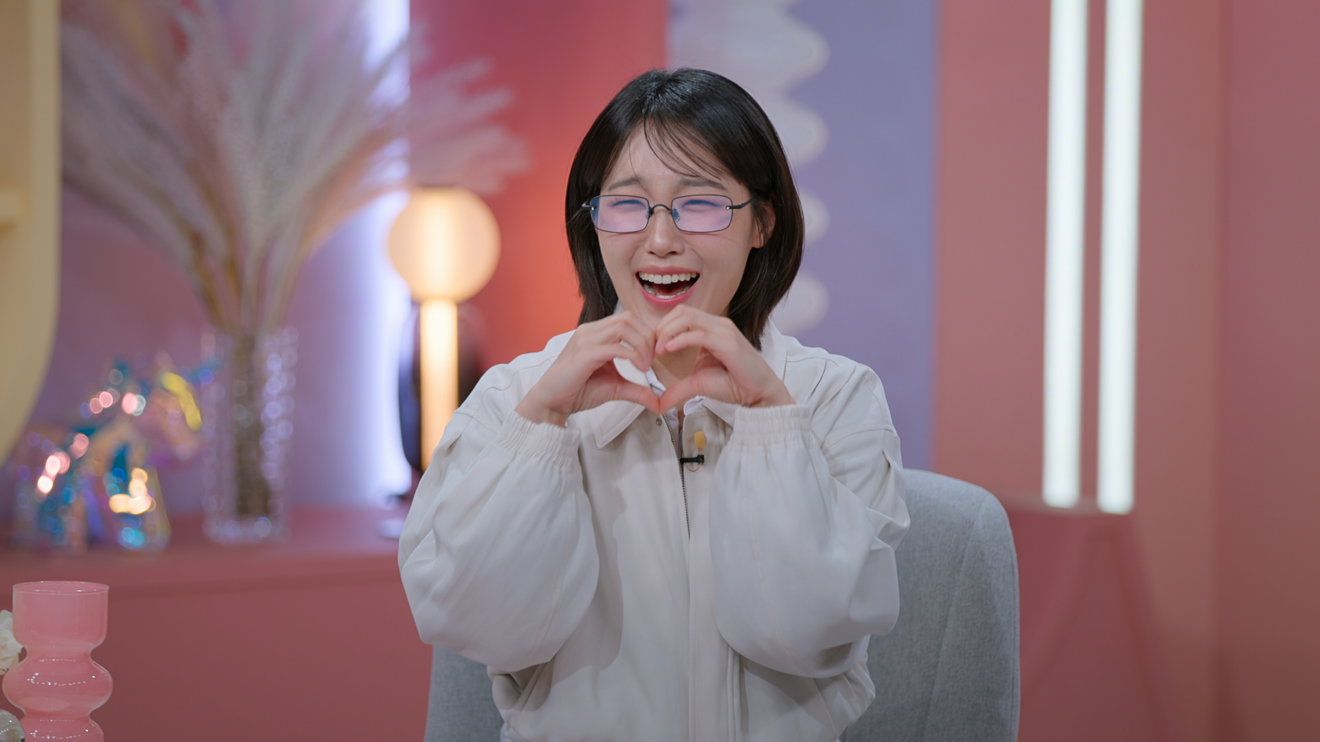 Co-host Lee Eun-ji makes a heart with her hands while wearing glasses and a gray jacket. She sits in a light-pink lounge, in a still from Netflix's 'Better Late Than Single.'
