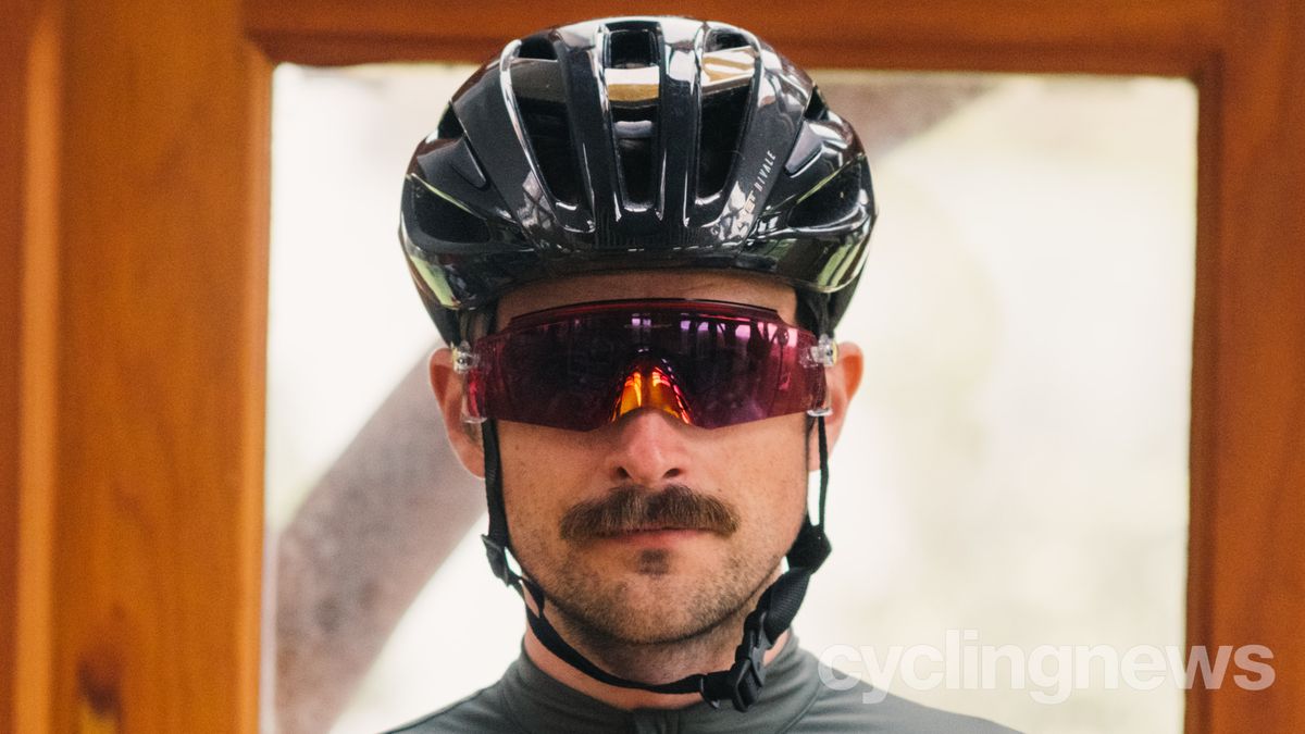 A white man with a moustache wears a red pair of Oakley Kato glasses under a black helmet