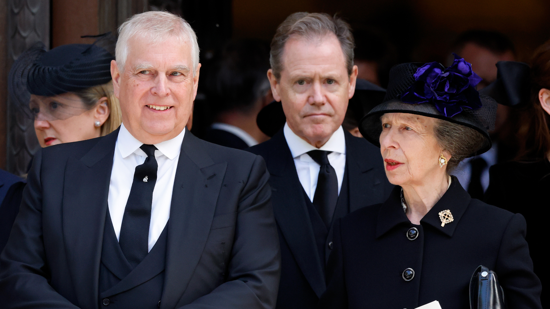 Prince Andrew wears a suit and has gray hair and smirks while his sister Princess Anne wears a black coat and hat and has red lipstick on