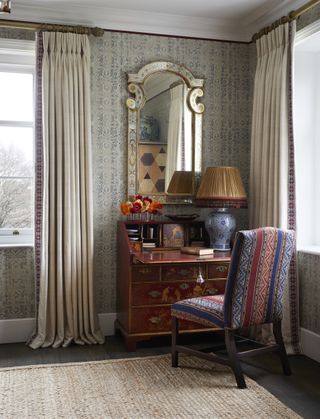 The corner of a room with patterned wallpaper with a dresser, an antique mirror, and a patterned chair