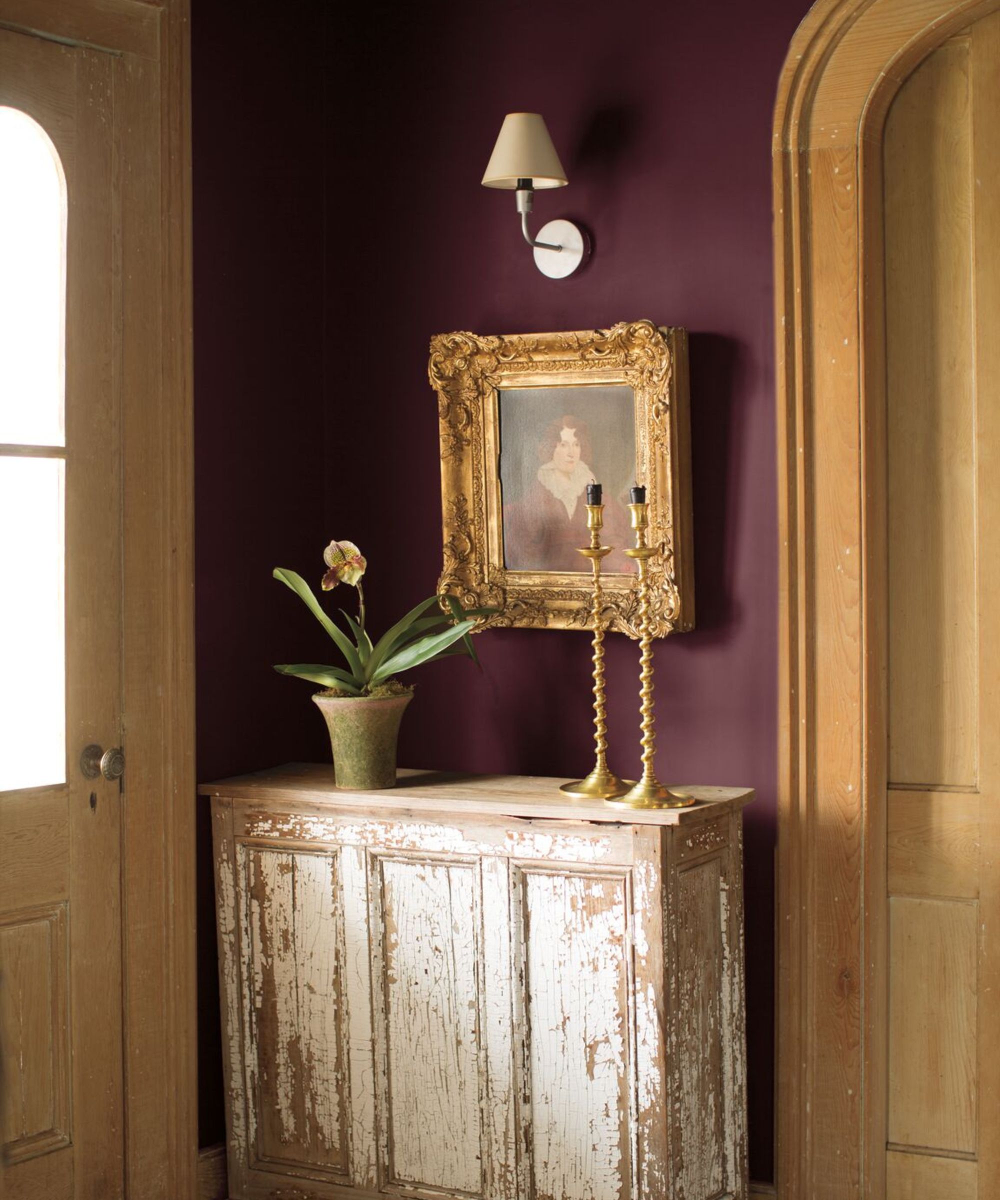 Entryway painted in Benjamin Moore's New London Burgundy, a distressed console table with gold candlesticks, a potted plant, and an antique painting, and a sconce above, and arched wooden doors.