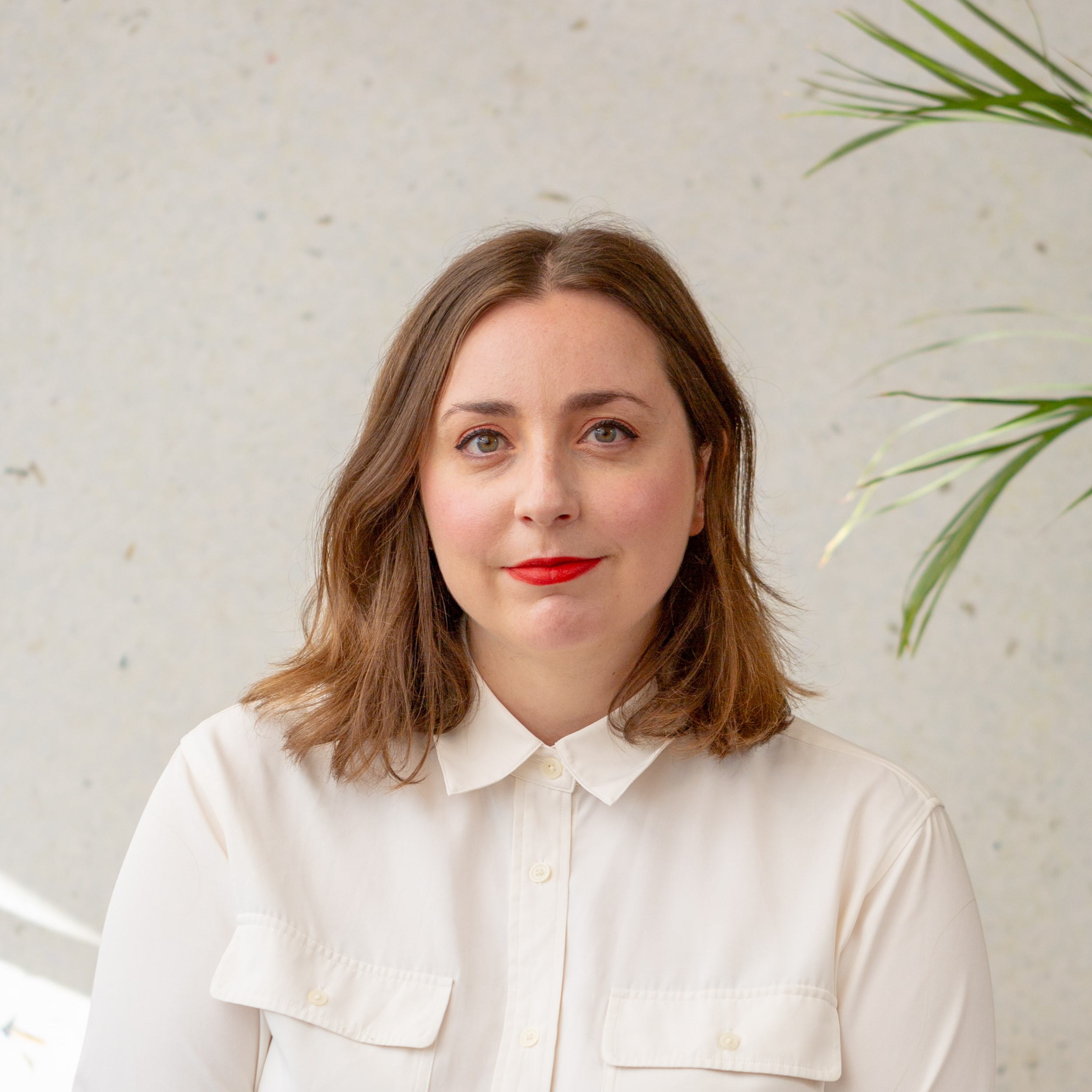 Lea Berger wears a white button-down shirt and red lipstick, posing for a headshot against a neutral, textured wall with a green plant in the corner.