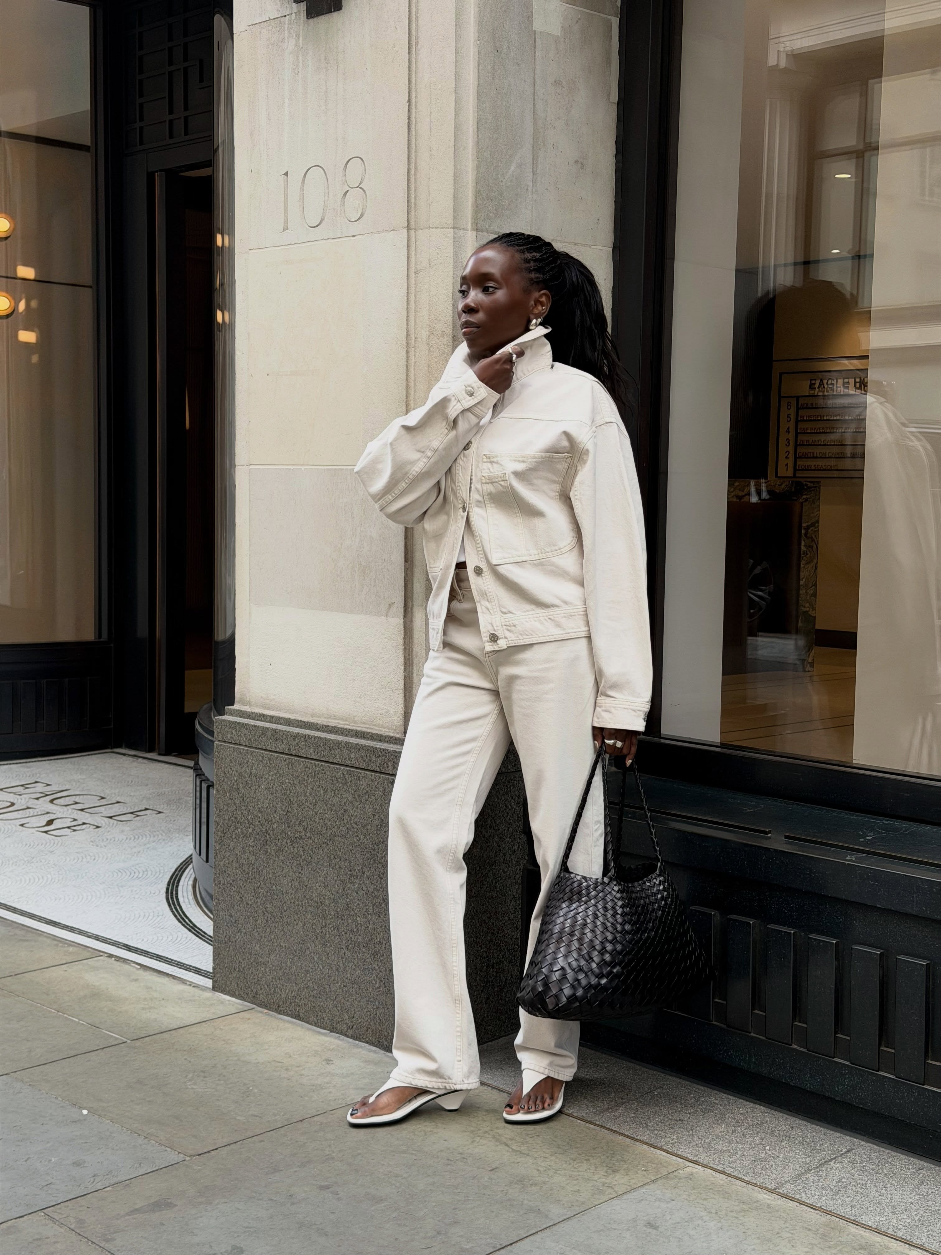 DanielleJinadu wears June Outfit Ideas 2026. She wears a white denim jacket, white tank, and white barrel leg jeans. She wears it with white kitten heel sandals and a black Dragon Diffusion bag.