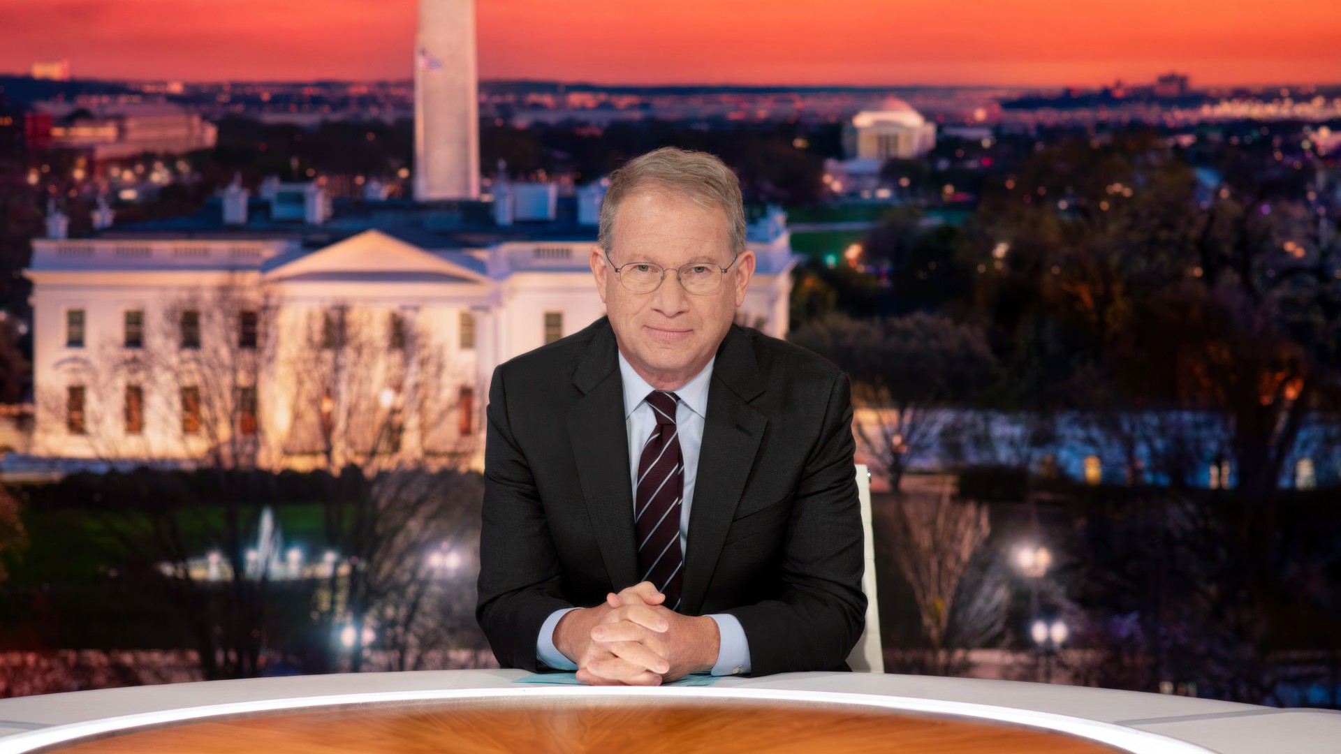 A man in glasses and a dark suit sits at a curved news desk, with the White House and Washington Monument visible at dusk behind him.