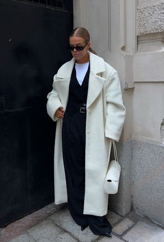 The winter capsule wardrobe item, a cashmere sweater, is shown in a photo of a woman standing outside wearing a long white coat over a white t-shirt layered under a v-neck sweater worn with a belt and black wide-leg trousers with pointed pumps, and white shoulder bag