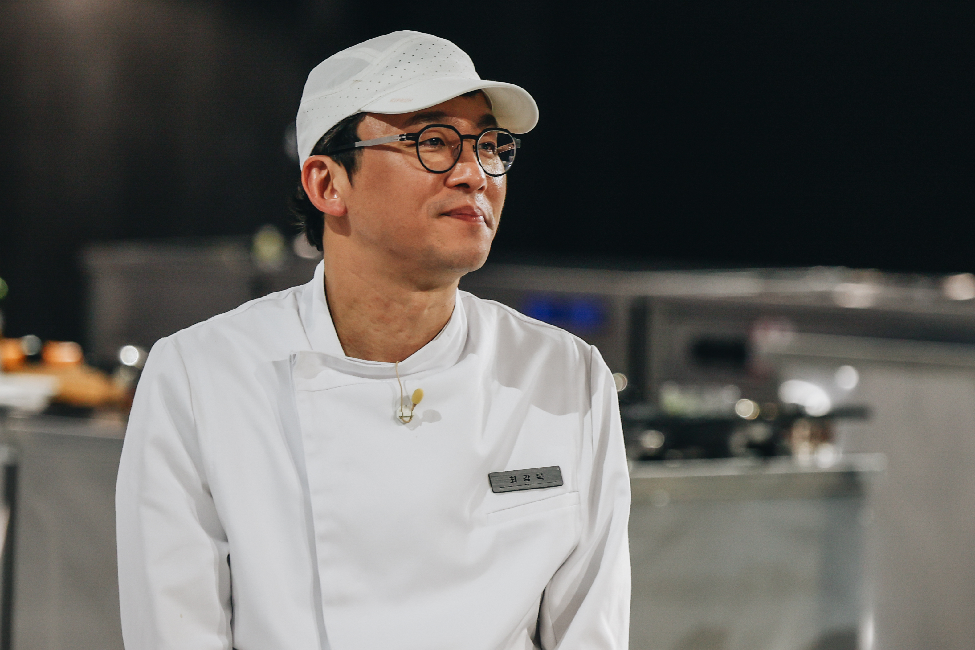 culinary class wars season 2 winner choi Kang-rok wearing a white baseball hat and white chef's coat smiling in a kitchen in a still from the reality show