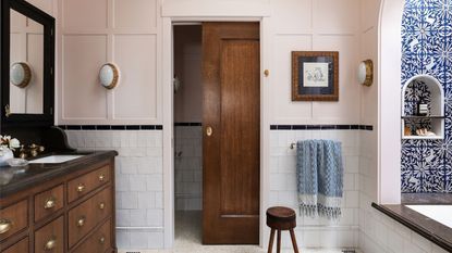 A bathroom with peach pink panelled walls, white tiles on the lower walls, a large wooden vanity with dark brown countertops, double sinks and mirrors, and a wooden sliding door. 