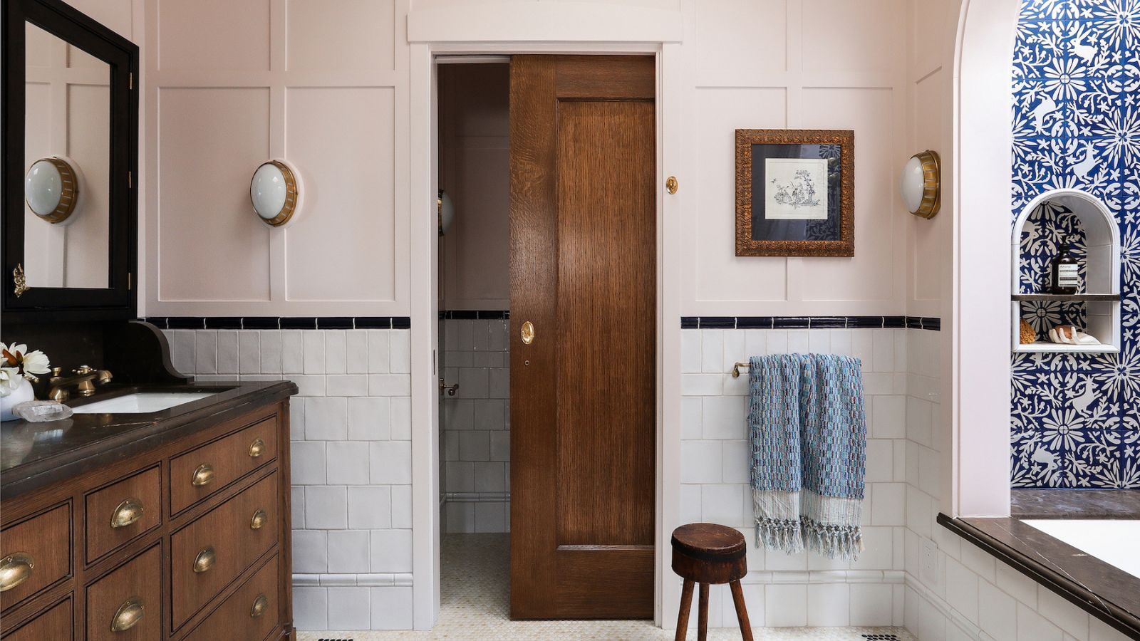 A bathroom with peach pink panelled walls, white tiles on the lower walls, a large wooden vanity with dark brown countertops, double sinks and mirrors, and a wooden sliding door. 