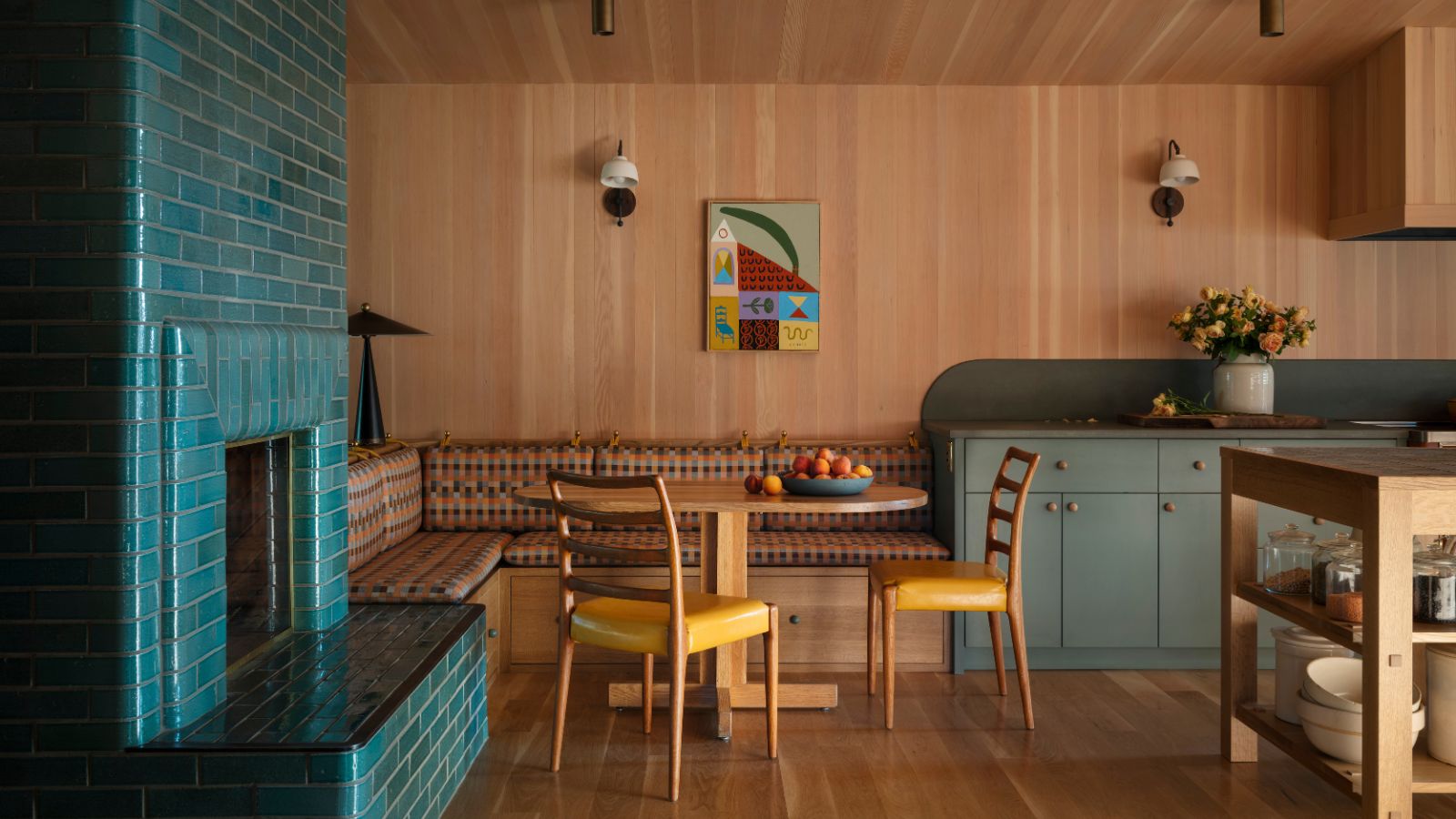 A wood-clad open-plan kitchen with wood floors, gray-green cabinets, banquette seating with a wood dining table and two mustard chairs. A teal brick fireplace. 