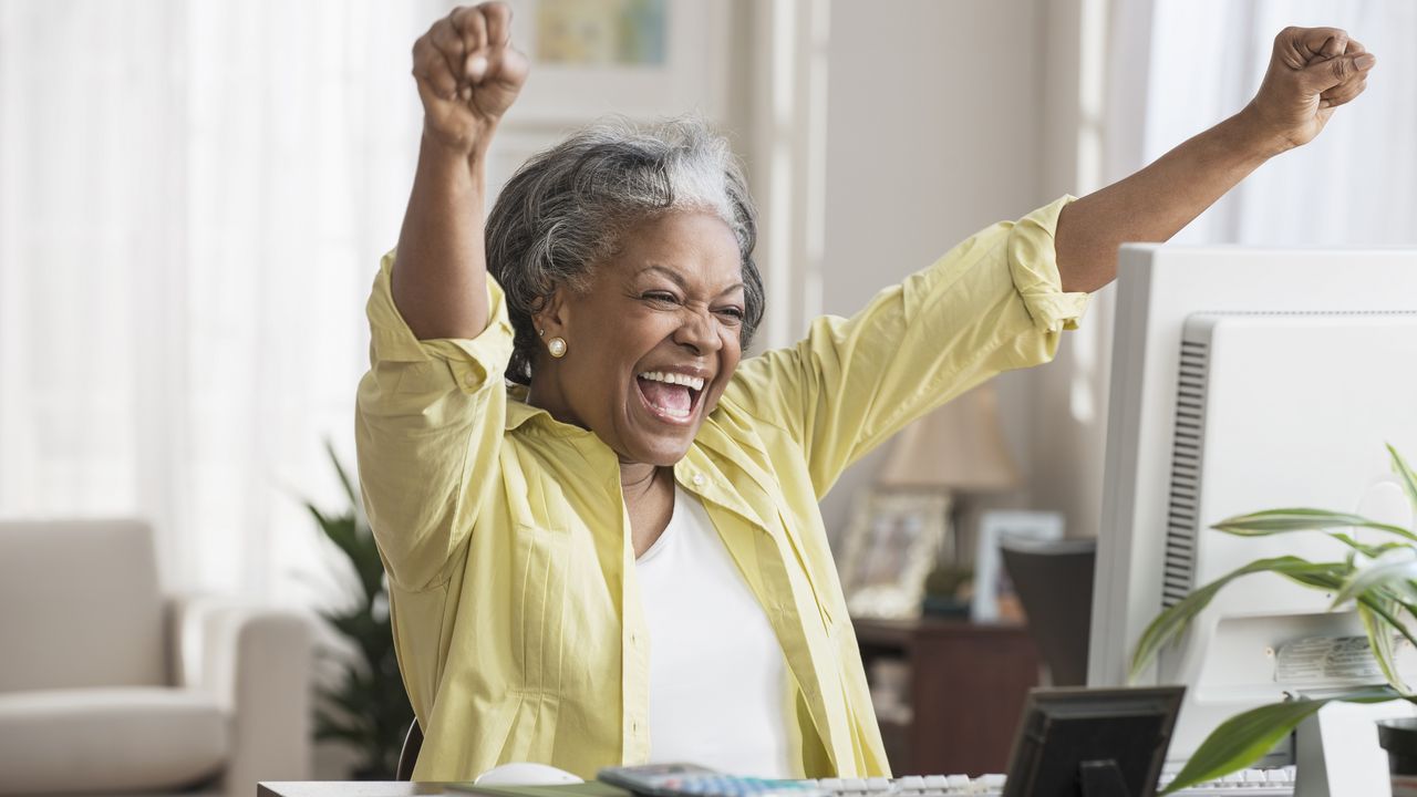 An older woman celebrates while looking at her computer screen at her desk.