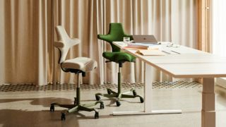 Two H&Aring;G Capisco ergonomic office chairs, one in cream fabric and one in forest green, are positioned at a white sit-stand desk in a sunlit room with beige curtains.
