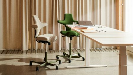 Two HÅG Capisco ergonomic office chairs, one in cream fabric and one in forest green, are positioned at a white sit-stand desk in a sunlit room with beige curtains.