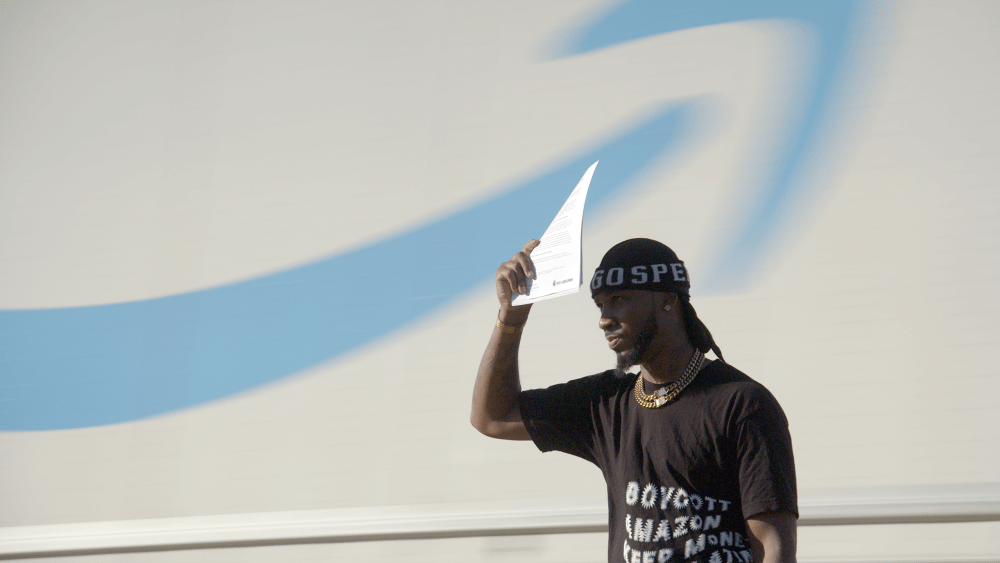 a man wearing a black printed shirt and cap holds up a paper to block the sun, while standing in front of a white wall with the amazon smile logo