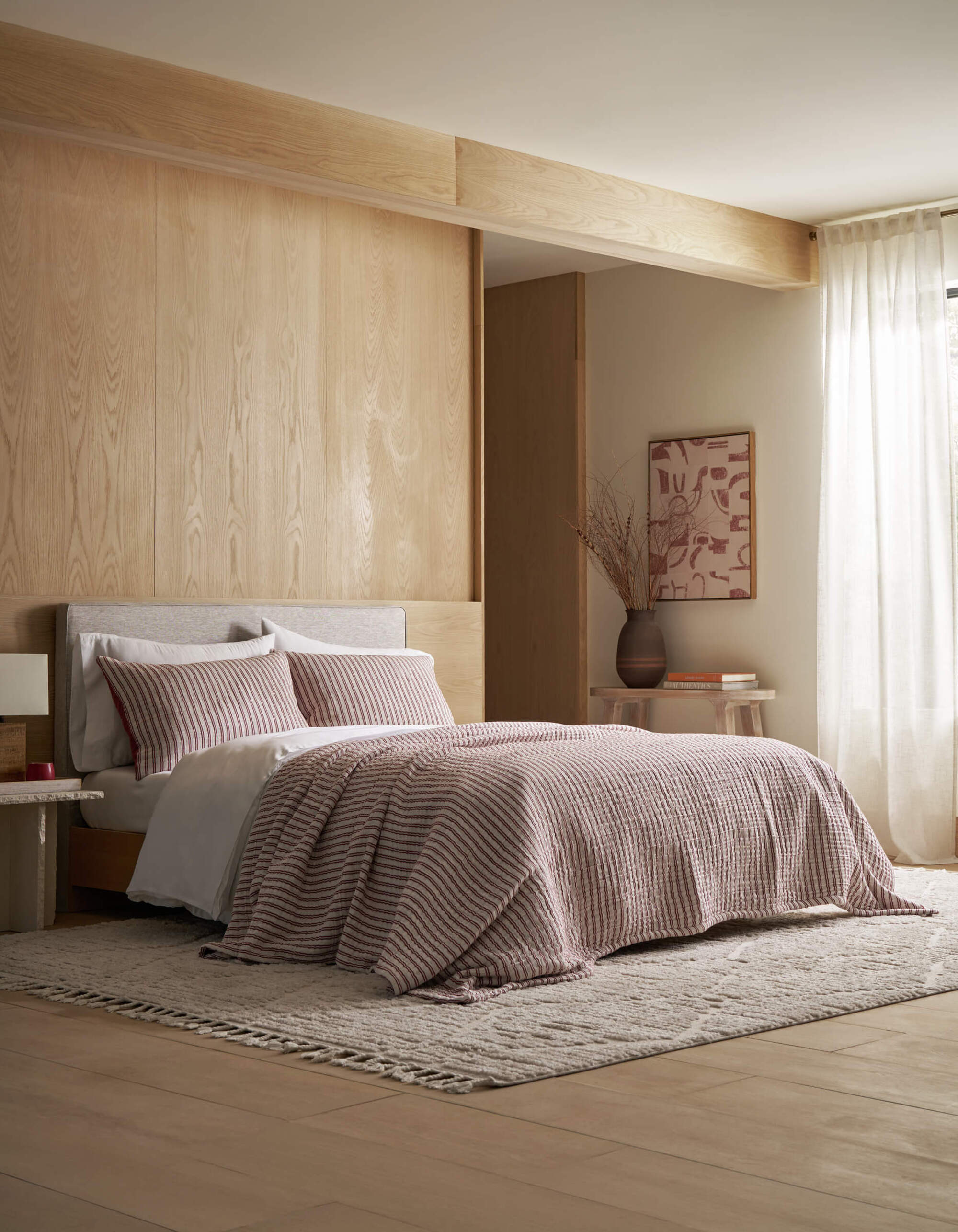 Image of a bed in a minimalist, light wood room. There is an off-white rug under the bed, and the bed has red and white striped bedding.