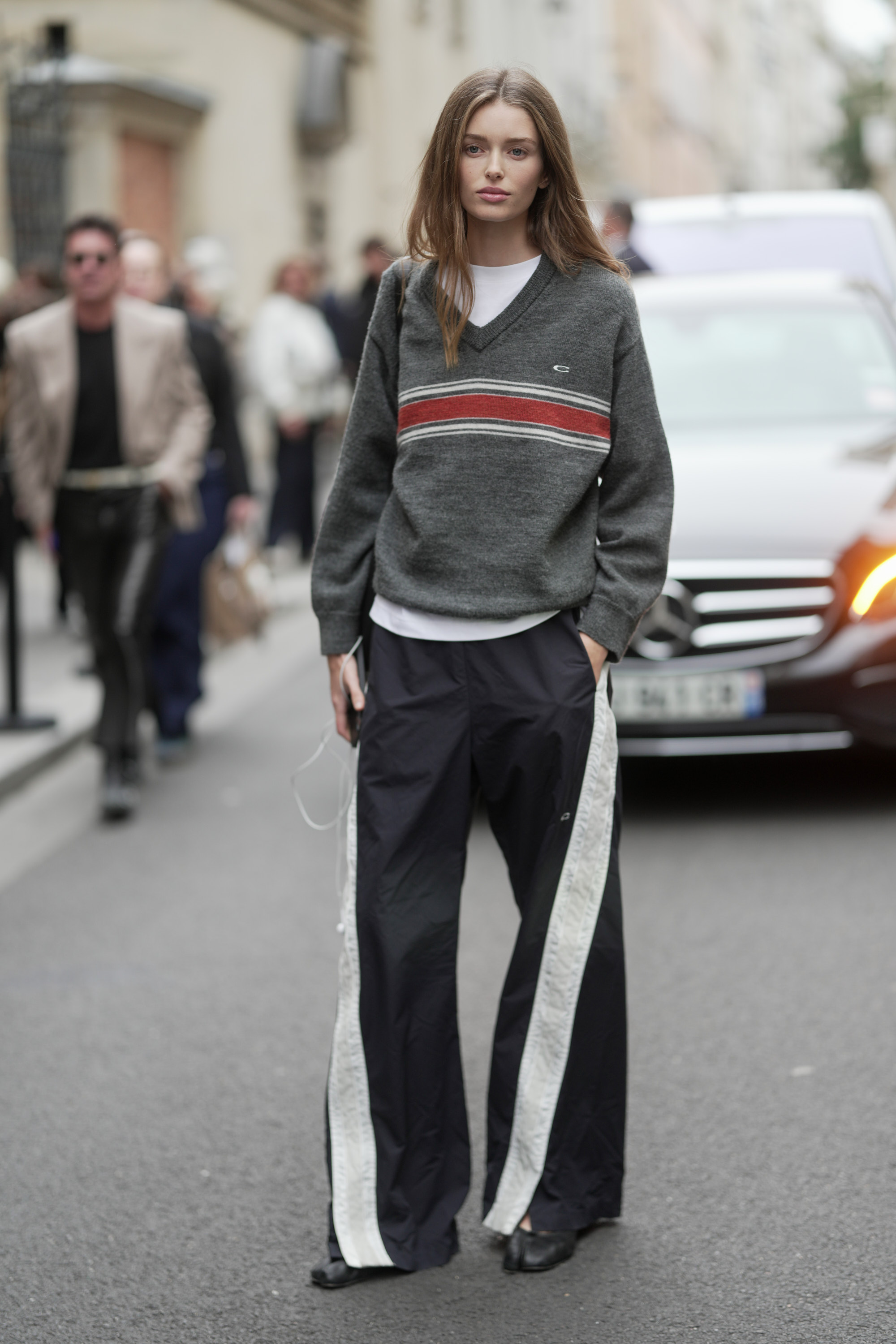 PARIS, FRANCE - OCTOBER 06: Stella Hanan is seen wearing loose, straight brown hair, a white crew-neck T-shirt, a grey V-neck sweater with red and white stripes, a black crossbody bag, wide-leg black track pants with white side panels, and black pointed-toe shoes outside of Sacai during the Womenswear Spring Summer 2026 as part of Paris Fashion Week on October 06, 2025 in Paris, France. (Photo by 305pics/Getty Images)