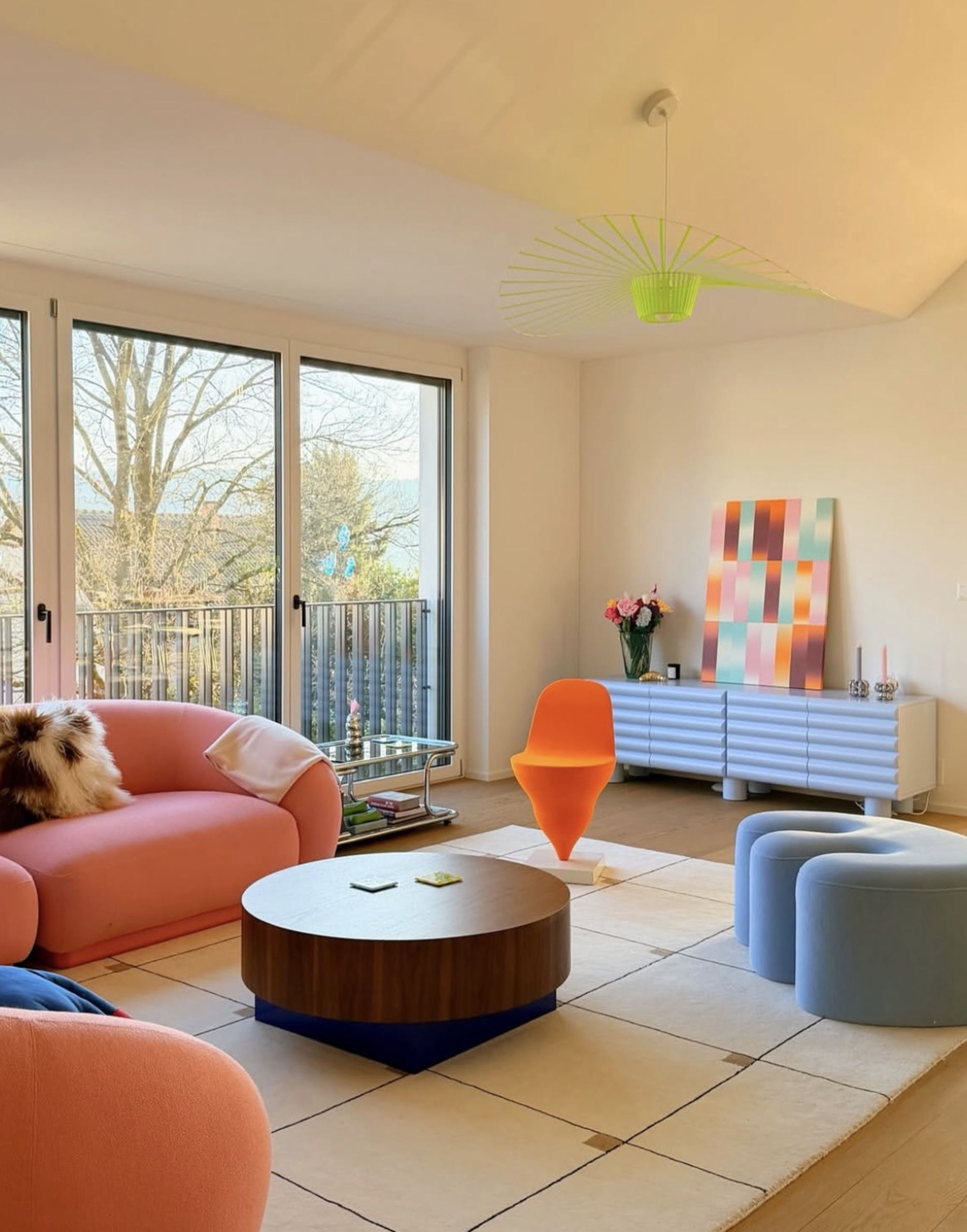 Image of a modern, bright living room with floor to ceiling glass doors on one wall. There is a pink sofa, a blue pouffe, and a blue console with a gradient painting on it.