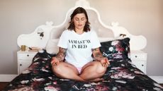 Woman sits cross-legged on a bed spead wearing a white T-shirt that reads NAMAST&rsquo;AY IN BED