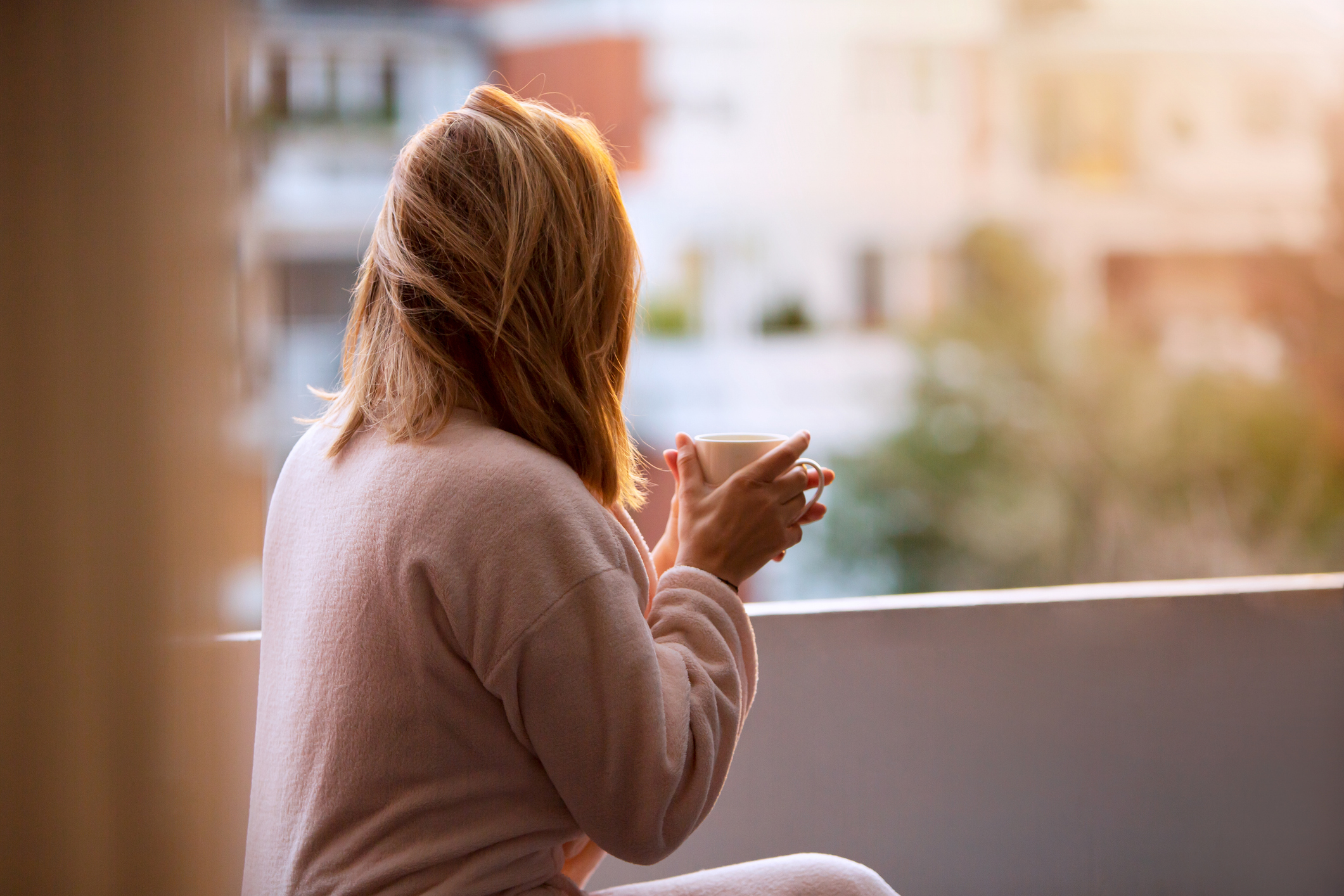 A woman drinks a cup of coffee on a balcony as the sun rises, wearing a comfortable outfit.
