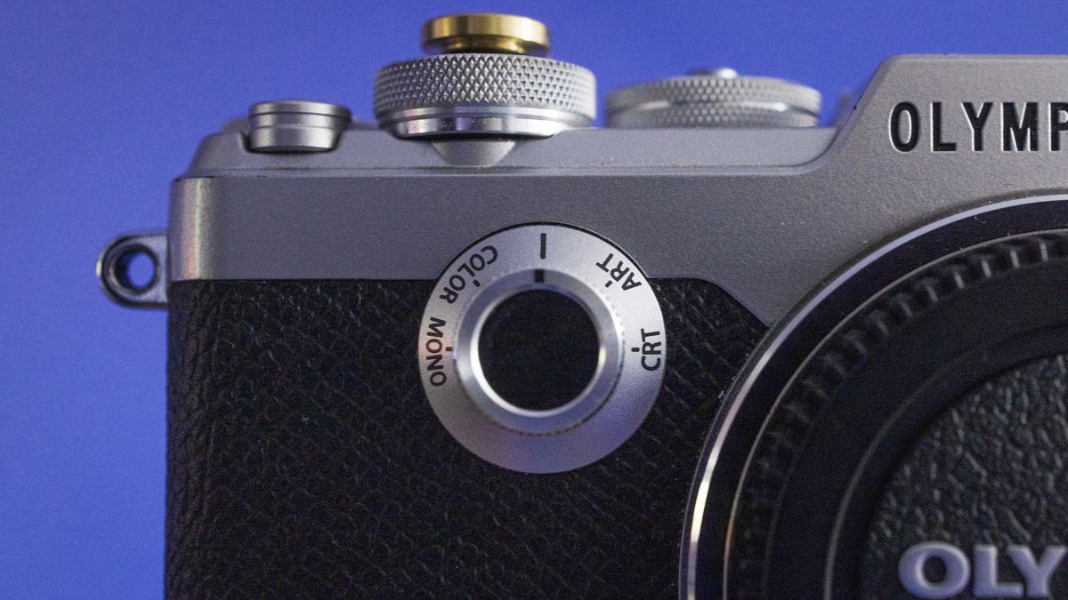 Close-up of an Olympus PEN-F camera body, against a purple background, focusing on the Creative Dial